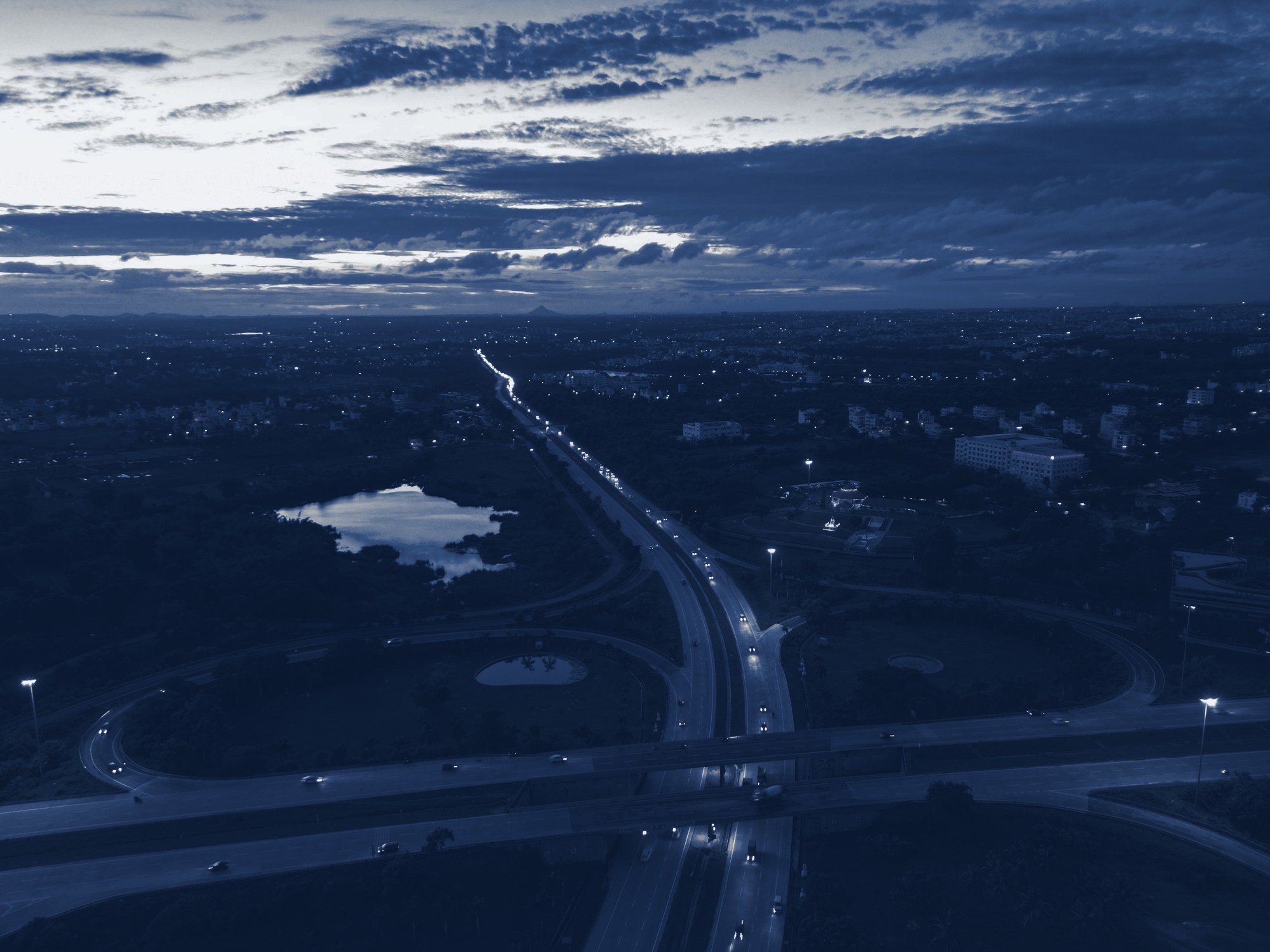 Aerial of a highway interchange at dusk with car light trails and a city spreading to the horizon, tinted navy