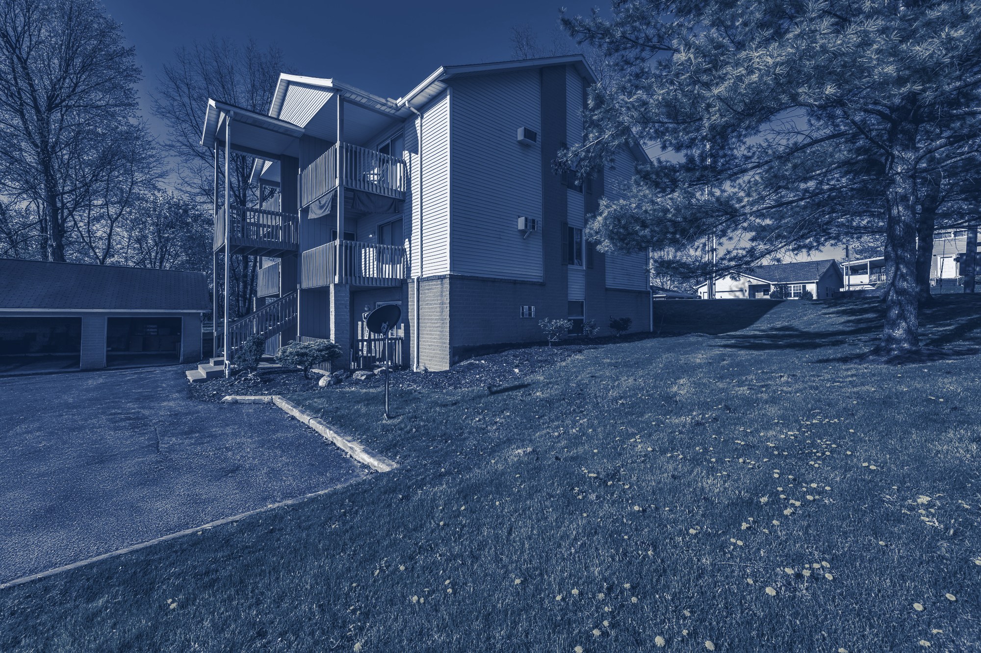 Three-story garden apartment building with wooden balconies beside a grassy lawn and trees, tinted navy