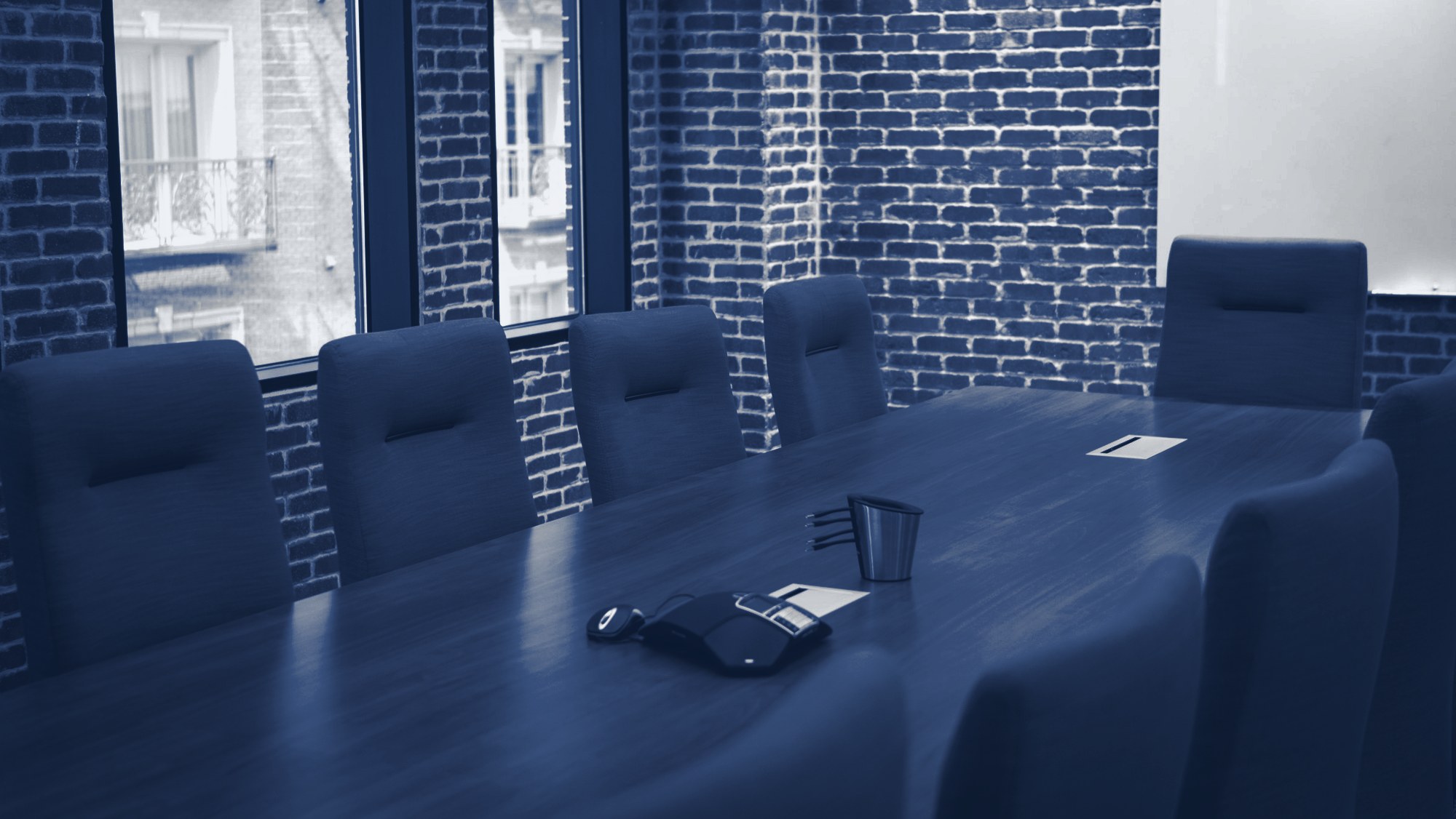 Empty boardroom: long wooden conference table, upholstered chairs, brick walls, conference phone and a sheet of paper on the table, tinted navy